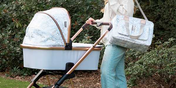 A mother walking her baby in a My Babiie MB500 Dani Grey Marble travel system in a garden.